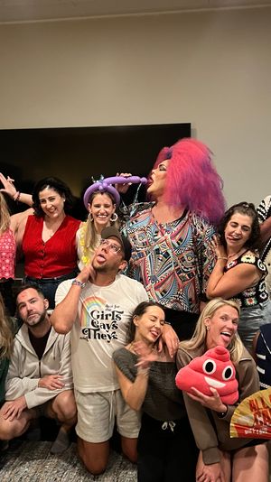 Lively at-home party group photo in a living room with friends and a drag performer in a bright pink wig, balloon hat, plush emoji and fan, all making goofy faces.