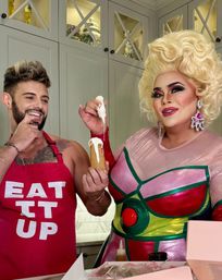 Drag performer in a colorful metallic costume and big blonde wig pipes white frosting onto a pastry cone while a smiling person in a red "EAT IT UP" apron watches in a bright home kitchen.