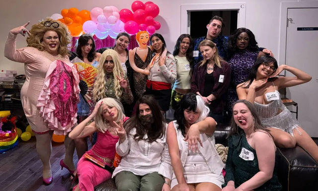 Playful group photo of adults in colorful costumes and drag posing and laughing at an indoor party with a pink-and-orange balloon arch and inflatable doll