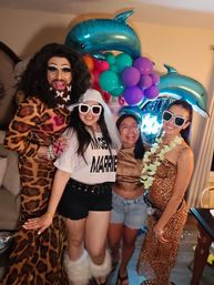 Four friends at a home bachelorette party posing in front of teal dolphin and colorful balloon decorations — a drag performer in leopard-print gown with butterfly beard, a veil-wearing bride-to-be in white tee and fluffy boots, and two women in festive sunglasses and lei.