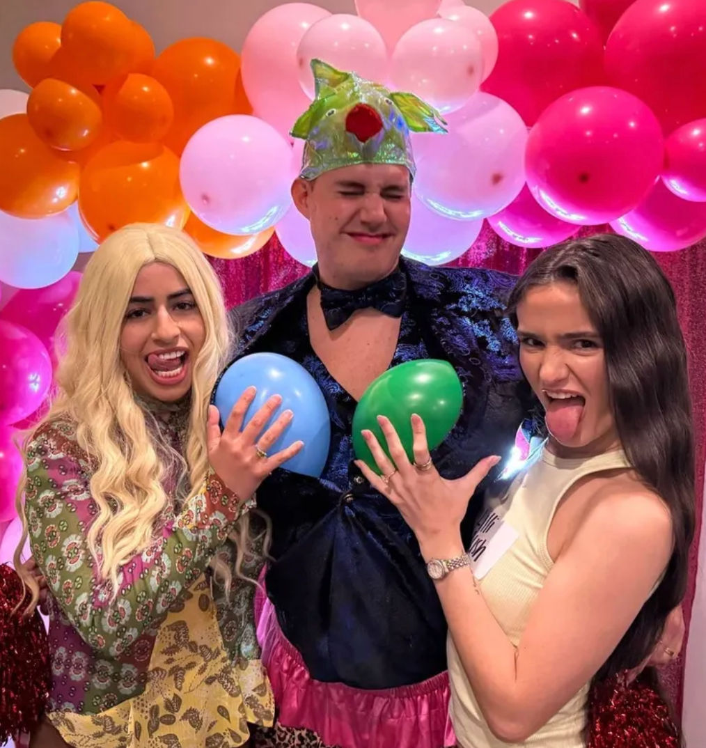 Three friends at an indoor celebration posing in front of a pink and orange balloon backdrop; person in the center wears a green crown and holds blue and green balloons to their chest while two friends laugh and stick out their tongues.