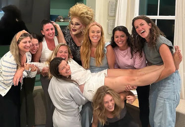Smiling group of women in a cozy living room holding a friend horizontally during a lively friends’ celebration, with a glamorous drag performer in a sparkly dress and big blonde wig joining the fun.