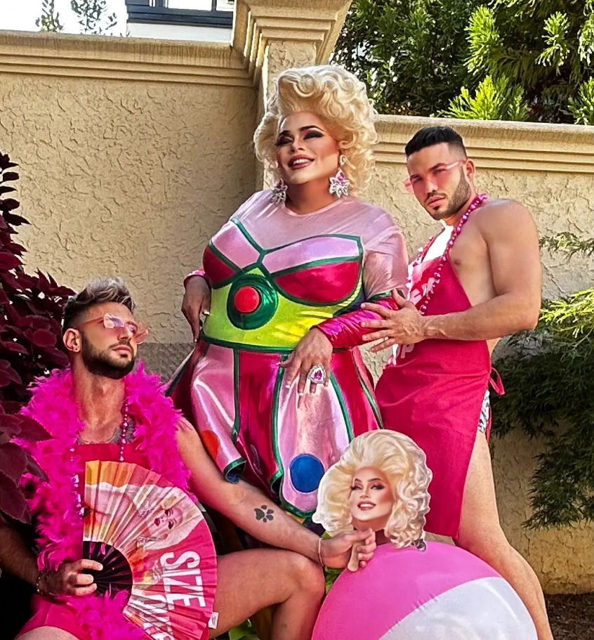 Glitzy outdoor patio scene with a glamorous drag performer in a metallic pink-and-green dress and blonde wig, flanked by two companions in pink outfits—one with a feather boa and fan, the other in a pink apron posing with a beach ball cutout; stucco wall and greenery in the background.
