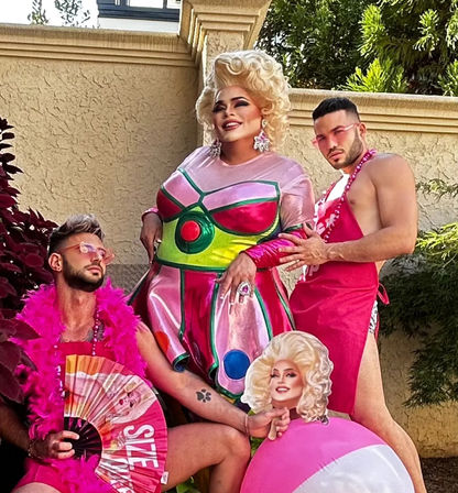 Glitzy outdoor patio scene with a glamorous drag performer in a metallic pink-and-green dress and blonde wig, flanked by two companions in pink outfits—one with a feather boa and fan, the other in a pink apron posing with a beach ball cutout; stucco wall and greenery in the background.