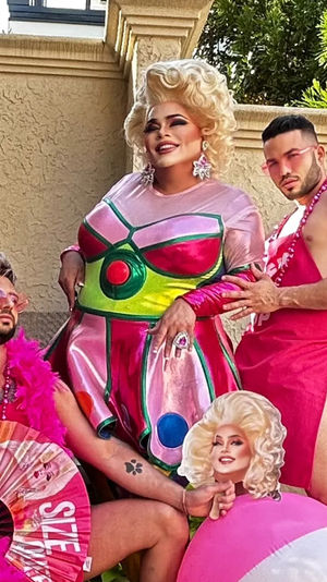 Glitzy outdoor patio scene with a glamorous drag performer in a metallic pink-and-green dress and blonde wig, flanked by two companions in pink outfits—one with a feather boa and fan, the other in a pink apron posing with a beach ball cutout; stucco wall and greenery in the background.