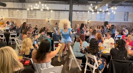 Drag performer in a blue sequined costume and big blonde wig parades through a lively indoor drag brunch as guests at tables cheer, clap and film in a brick-walled industrial event space with hanging lights.