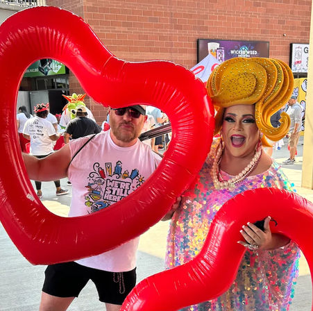 Two attendees framed by oversized red heart inflatables at an outdoor Pride festival — a man in sunglasses and a graphic tee beside a jubilant drag performer in a sparkling sequin dress and large yellow wig, brick wall and crowd behind them.