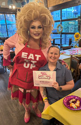 Drag performer in a big blonde wig and red apron reading "EAT IT UP" poses with a smiling person holding a "Winner" certificate at a colorful indoor restaurant celebration with sunflowers and decorated tables.