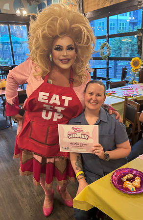 Drag performer in a big blonde wig and red apron reading "EAT IT UP" poses with a smiling person holding a "Winner" certificate at a colorful indoor restaurant celebration with sunflowers and decorated tables.