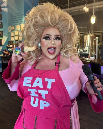 Energetic drag performer in a huge blonde wig and bold makeup wearing a bright pink apron reading “EAT IT UP,” holding a novelty cookie and a microphone inside a lively craft brewery with stainless steel tanks