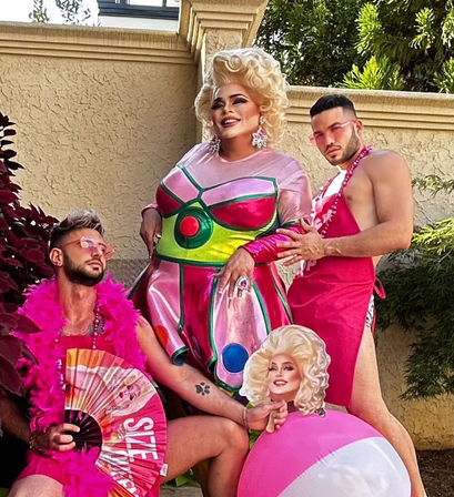Glamorous drag performer in a metallic pink-and-green dress and blonde wig poses on a sunny backyard patio against a stucco wall, flanked by two men in pink accessories (boa, sunglasses, apron), one holding a fan and the other a pink beach ball with a cutout portrait — playful summer party vibe.