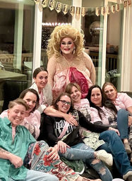 Birthday party at home: group of friends lounging on a living room couch with a glamorous drag performer in a pink sequined gown, 'Happy Birthday' banner overhead, dollar bills and matching party shirts — fun celebratory group photo.