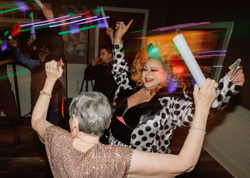 Drag performer in a polka-dot coat and big blonde wig dancing with partygoers on an indoor dance floor, colorful neon light trails, foam party stick and cash being waved