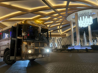Vintage rust-colored trolley bus parked under a glowing geometric hotel canopy at night beside an illuminated fountain and palm trees