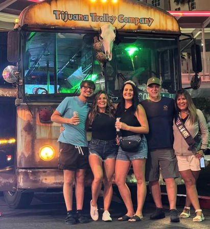 Five friends smiling and posing in front of a rustic party trolley with a skull ornament, neon-lit downtown nightlife scene.