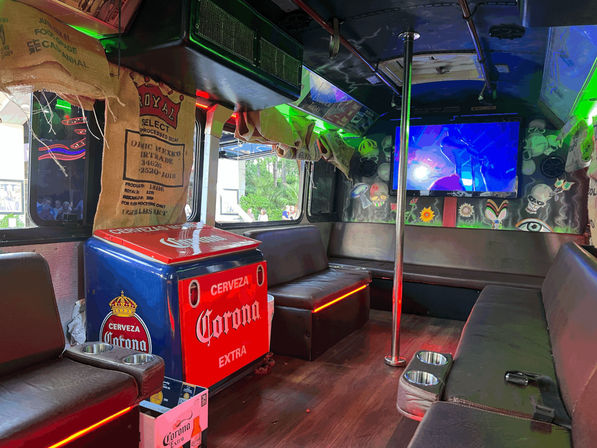 Vibrant party bus interior with leather bench seating, center dance pole, Corona-branded cooler, burlap sack curtains, neon LED accent lighting, a large wall-mounted TV showing colorful visuals and skulls and psychedelic mural art.