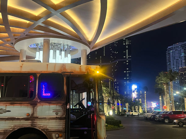 Rusty party bus with a blue neon sign reading LIVE NUDES parked under a glowing modern canopy at night, palm trees wrapped in string lights and high-rise buildings in the background.