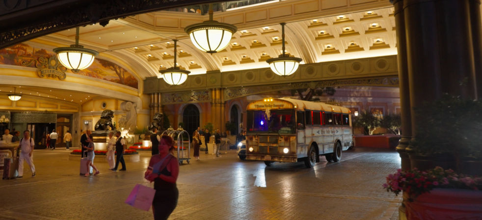 Nighttime luxury hotel entrance with grand hanging chandeliers, ornate coffered ceiling, travelers with luggage and a vintage shuttle bus.