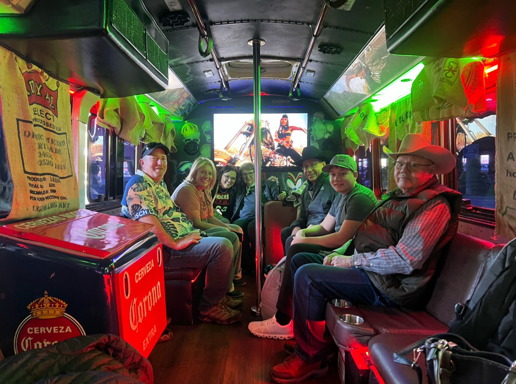 Smiling adults seated facing each other inside a neon-lit party bus interior, colorful red and green LED lights, beer cooler and burlap sack decor, central dance pole and a glowing screen showing a motorcycle image.