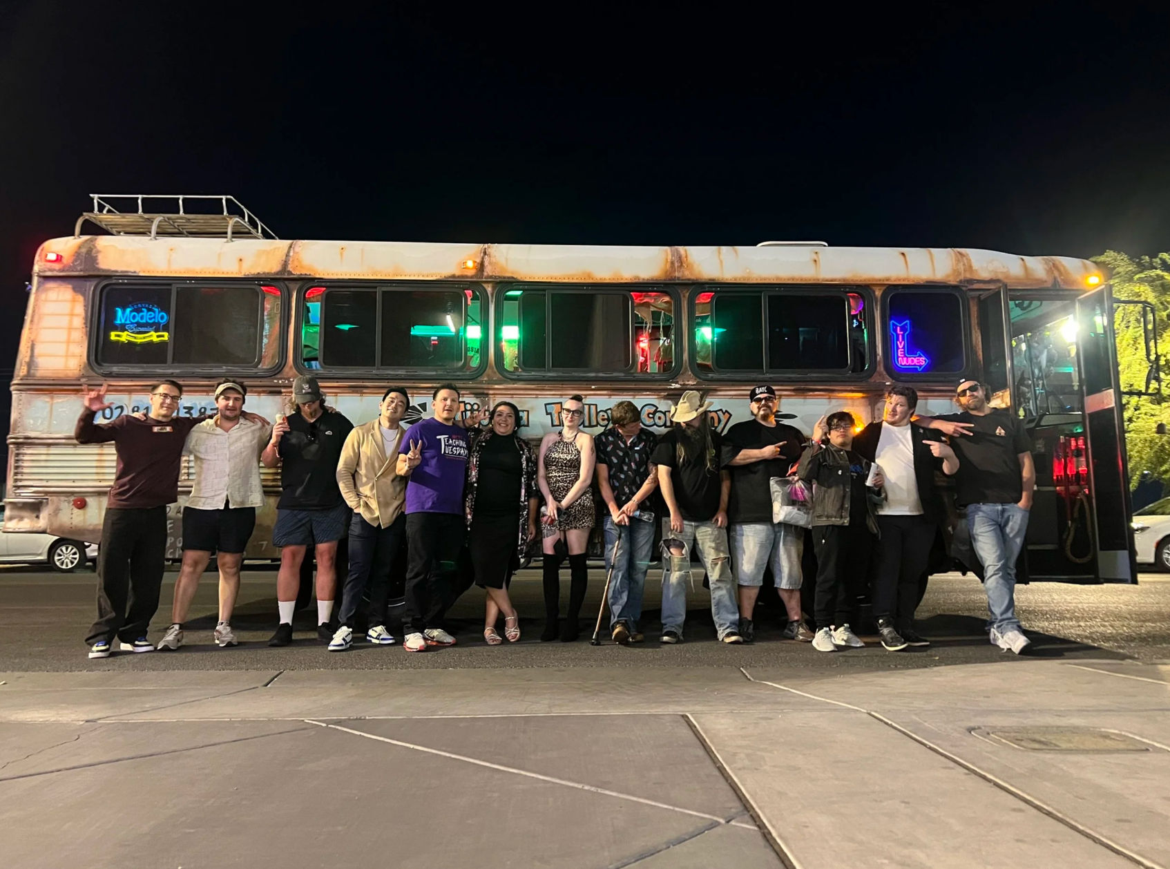 Group of people posing in front of a rusted retro party bus with neon signs, lit at night in a parking lot — lively nightlife group photo