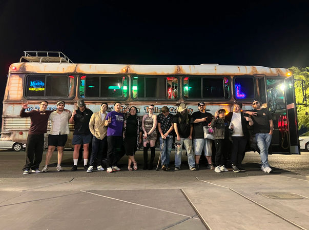 Group of people posing in front of a rusted retro party bus with neon signs, lit at night in a parking lot — lively nightlife group photo