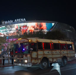 Night scene of a rusted vintage party bus with red interior lights driving past a brightly lit arena with a curved LED billboard on a city street
