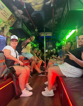 Smiling group of friends partying inside a neon-lit Las Vegas party bus with bench seating, a center dance pole, colorful green and red lights, and drinks in hand