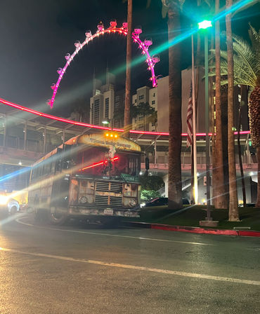 Nighttime Las Vegas street scene with a weathered trolley-style bus parked under a neon-lit pedestrian bridge, palm trees and a glowing observation wheel with illuminated pods above a hotel tower.