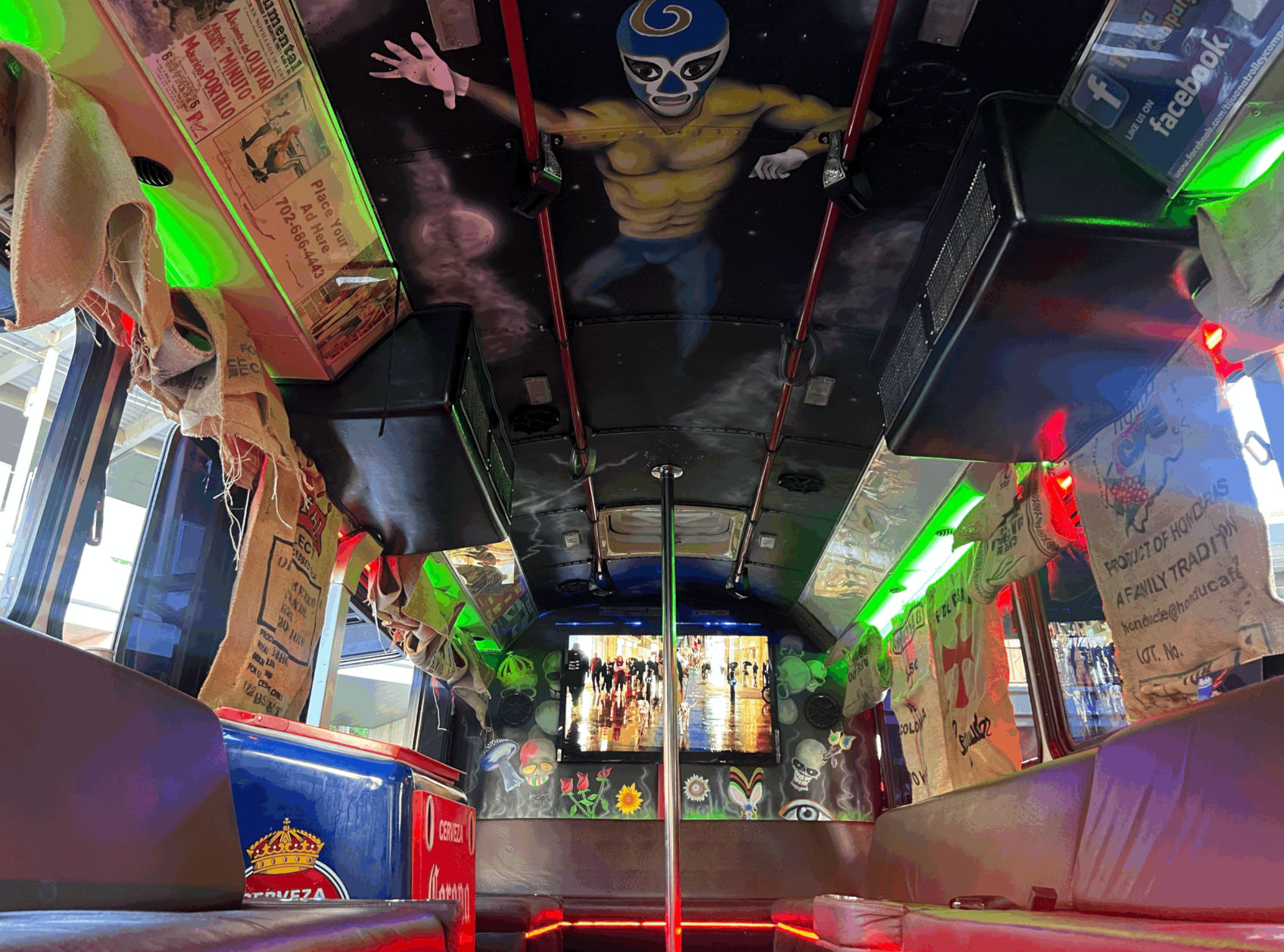 Vibrant party bus interior with cushioned bench seating, a chrome dance pole, ceiling mural of a masked luchador, burlap sack curtains and green and red LED accent lighting.
