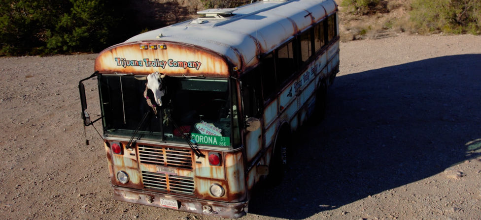 Rusty vintage trolley-style bus parked on gravel in an arid desert, cow skull mounted on the front, weathered paint and deep shadow.