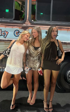 Three women in sparkly party outfits posing and laughing in front of a colorful party bus at night on a city street, holding drinks and wearing sandals for a lively night out.