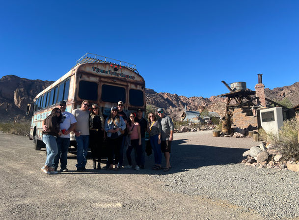 Smiling group posing by a rusted vintage trolley bus in a sunny desert ghost town with rocky hills and wooden mining structures under a clear blue sky