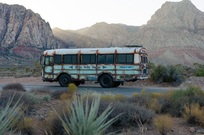 Rusty vintage trolley-style bus parked on a desert roadside among scrub and yucca plants, set against dramatic red‑rock mountains at sunrise.