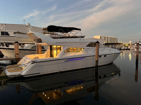 Sleek white motor yacht docked at a marina at dusk, warm cabin lights and blue accent lighting reflecting on calm waterfront