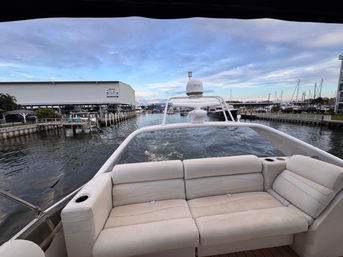 View from a yacht's aft lounge — white cushioned bench and cup holders overlooking a marina with docks, docked boats and the boat's wake under a pastel evening sky.