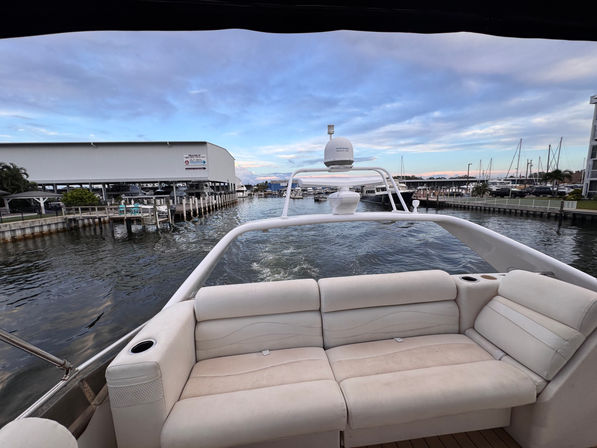 View from a yacht's aft lounge — white cushioned bench and cup holders overlooking a marina with docks, docked boats and the boat's wake under a pastel evening sky.