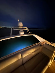 Night yacht deck with cushioned seating, blue underwater lights glowing in dark coastal waters and a radar dome under a cloudy sky