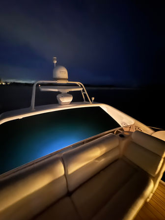 Nighttime view from a yacht's illuminated aft deck with cushioned seating in the foreground and blue underwater lights glowing off the transom beneath a dark coastal sky.