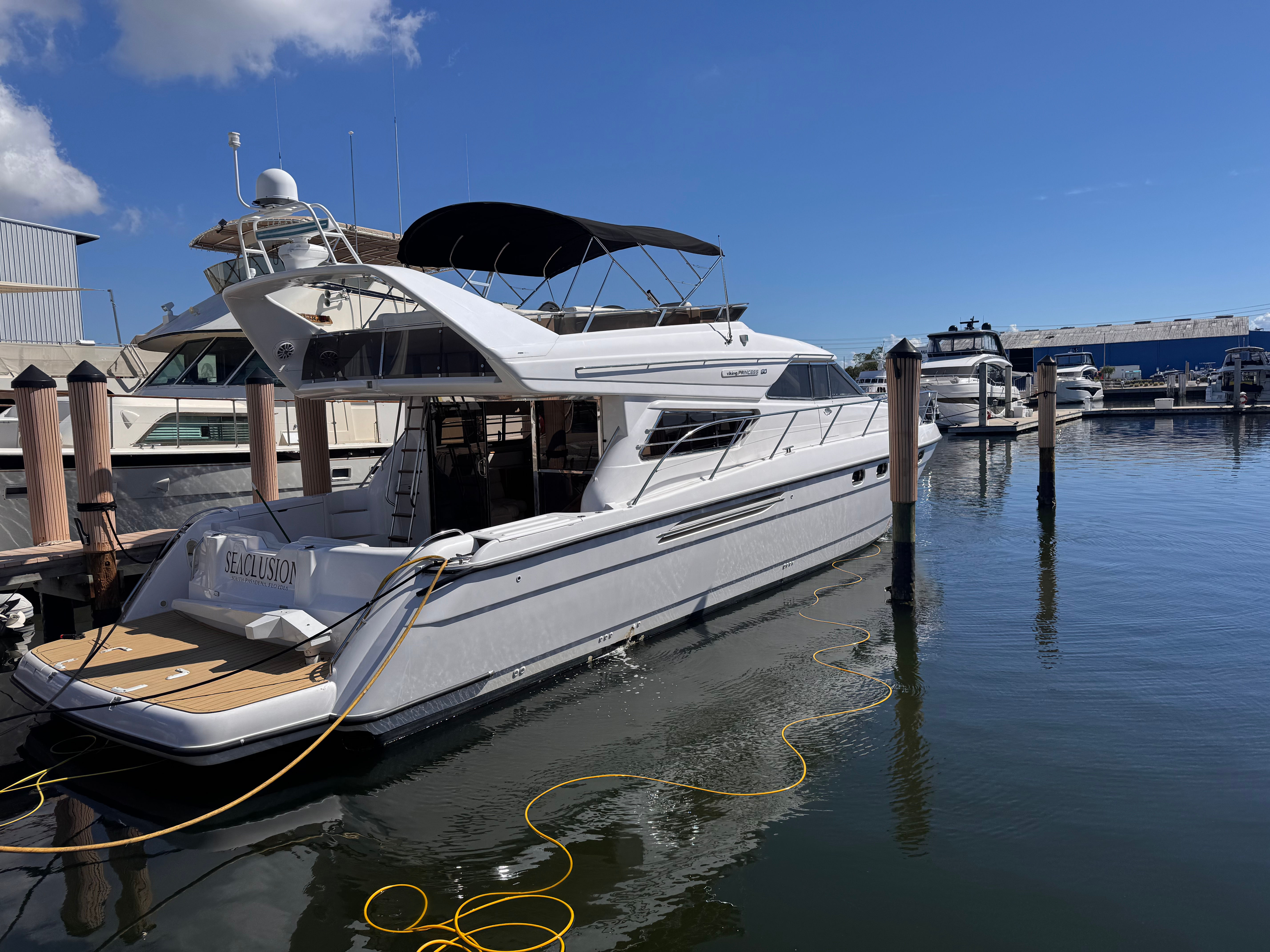 White luxury motor yacht moored at a sunny marina, docked by wooden pilings with a black bimini top, yellow power cable trailing through calm reflective water and other boats in the background.