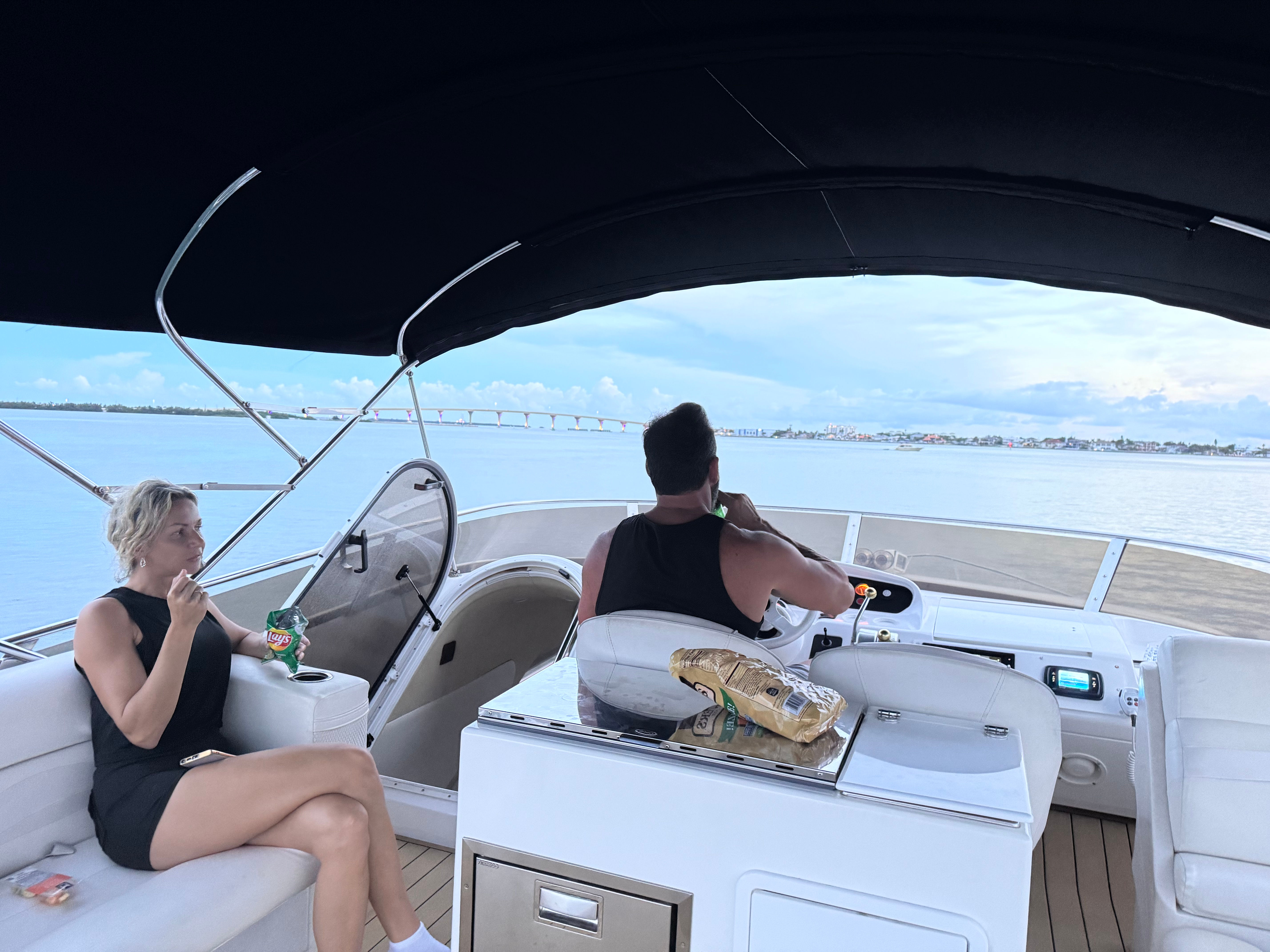 Two people relaxing on a covered motorboat — a man at the helm sipping a drink and a woman snacking on chips on cushioned seats, calm blue bay with a low bridge and distant coastal shoreline under a cloudy sky.