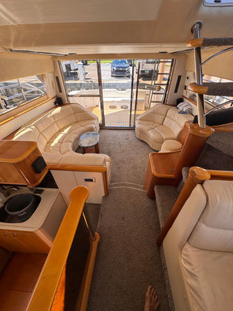 Sunny yacht salon with curved beige leather sofas, round marble-top coffee table, carpeted steps and spiral stainless handrail, sliding glass doors opening to the aft deck and marina