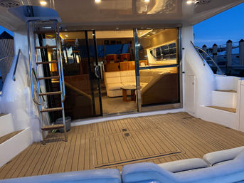 Luxury yacht aft deck at a marina at dusk with teak-style deck, stainless ladder to flybridge, sliding glass doors opening to a warm leather salon with curved sofa and coffee table.
