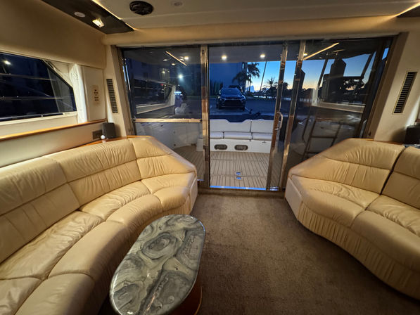 Luxury yacht salon interior with curved beige leather sofas and marble-effect coffee table, sliding glass doors opening to a teak aft deck and marina at sunset with silhouetted palm trees.