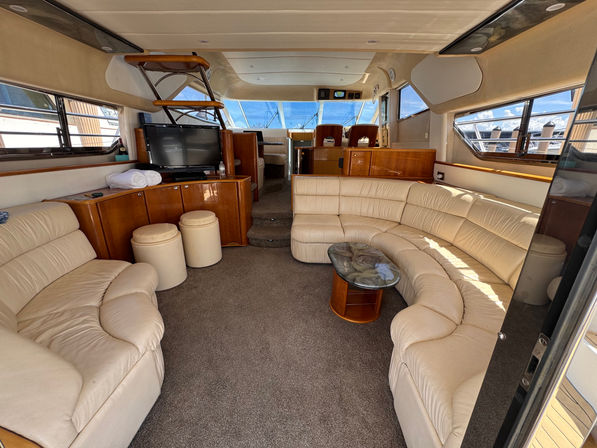 Sunlit luxury motor yacht salon with curved beige leather seating, wood veneer cabinets, glass-top coffee table, flat-screen TV, carpeted floor and steps leading to the forward helm with twin captain chairs and marina views through wide windows.