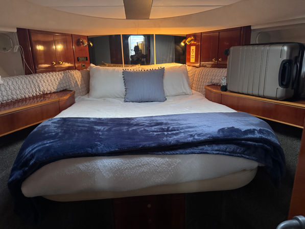 Cozy yacht stateroom with a center queen bed dressed in white linens, navy throw and a blue accent pillow, warm wood cabinetry, built-in reading lamps and a silver suitcase on a side shelf.