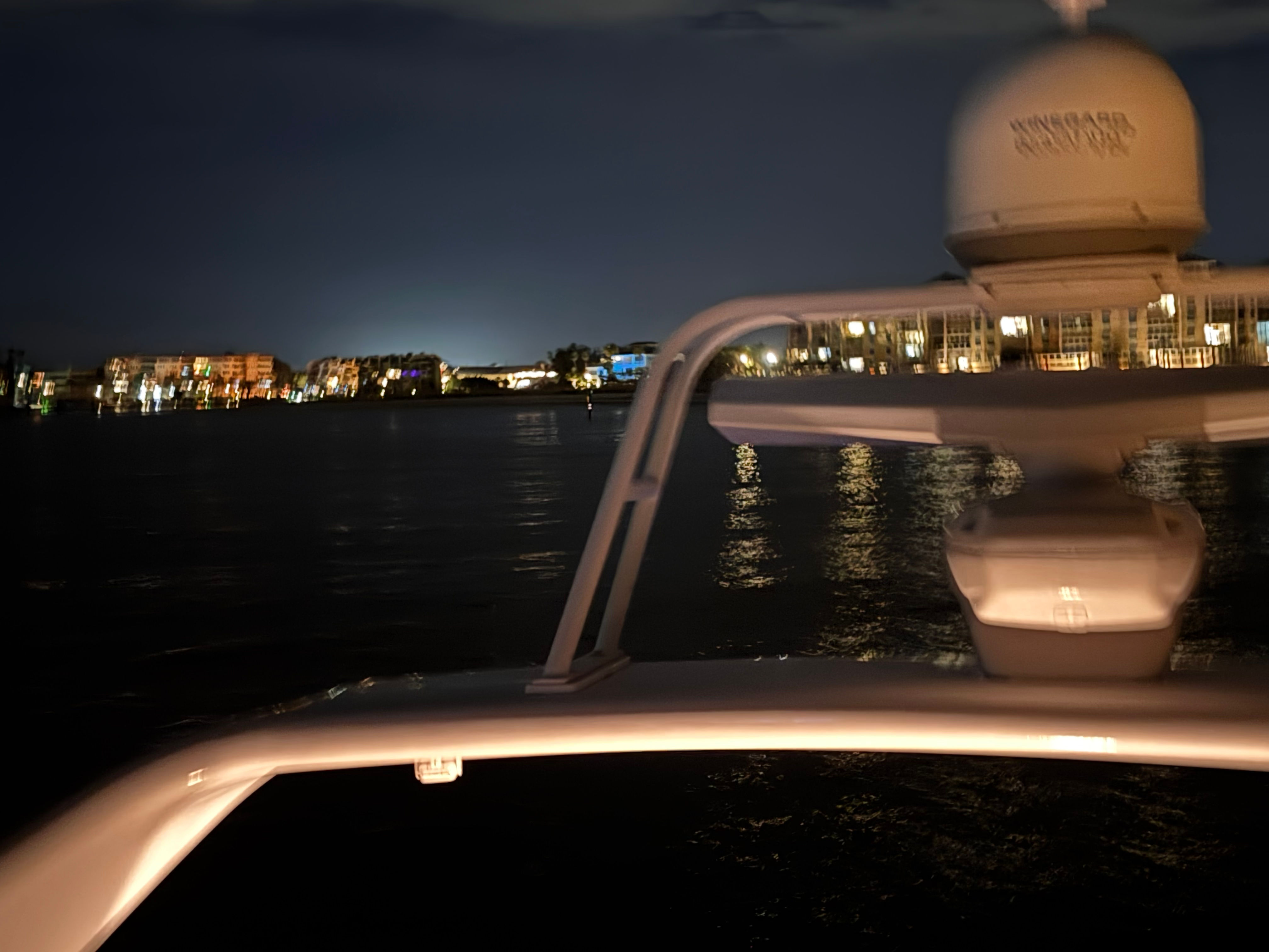 Nighttime waterfront view from a boat with the illuminated radar arch and bow in the foreground and dark water reflecting colorful marina and shoreline lights in the distance.