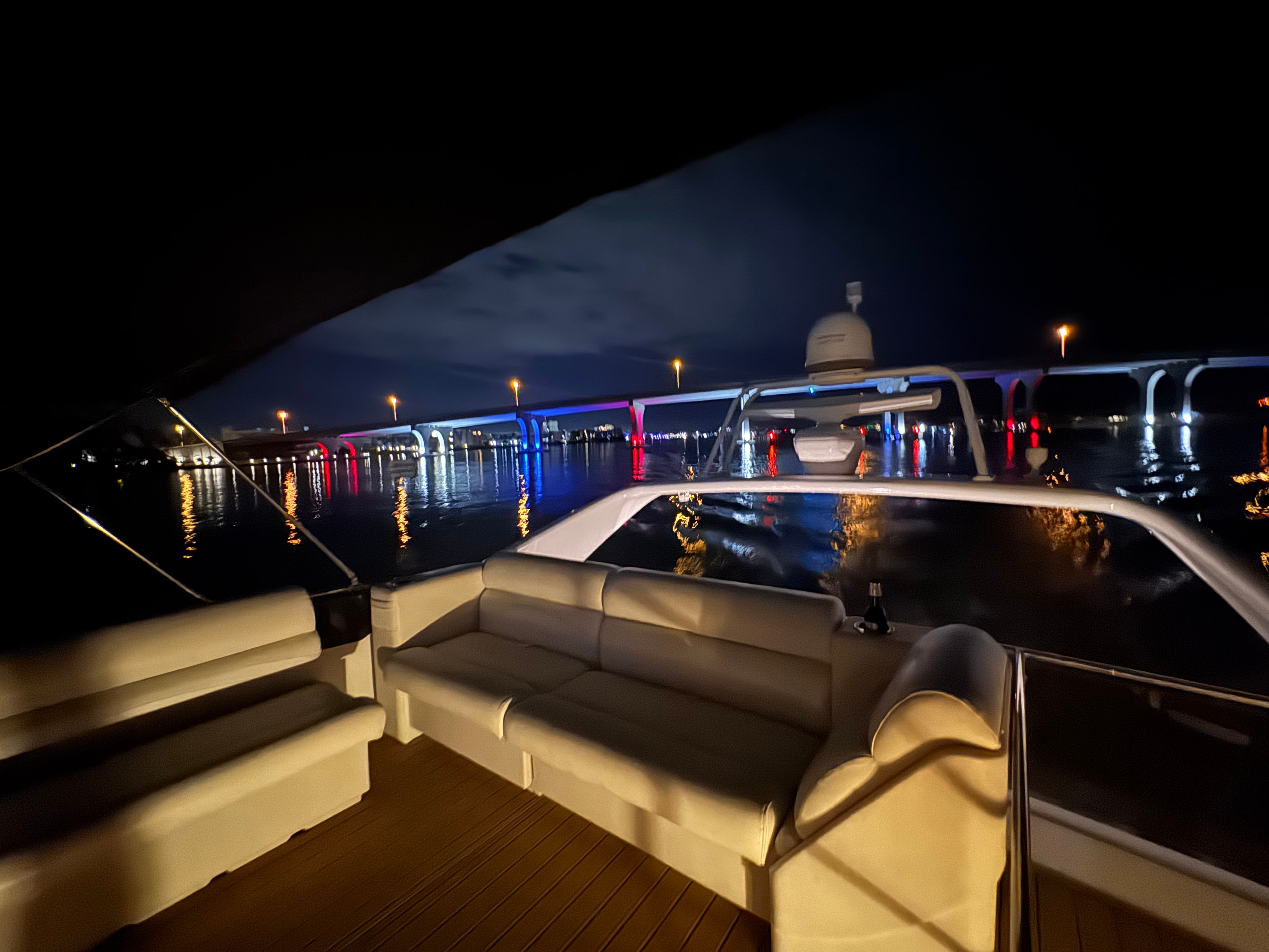 Nighttime yacht deck with white leather seating, looking out over calm water toward an illuminated arched bridge lit in red, white and blue with colorful reflections and city lights under a cloudy sky.