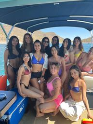 Group of smiling women in colorful swimsuits posing on a covered pontoon boat over turquoise water, with cactus-studded desert hills and clear blue sky in the background.