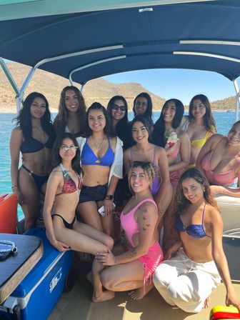 Group of smiling women in colorful swimsuits posing on a covered pontoon boat over turquoise water, with cactus-studded desert hills and clear blue sky in the background.
