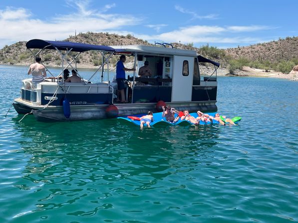 Pontoon boat anchored on a sunny desert lake with friends lounging on colorful foam floats in clear turquoise water near a rocky shoreline and blue sky.
